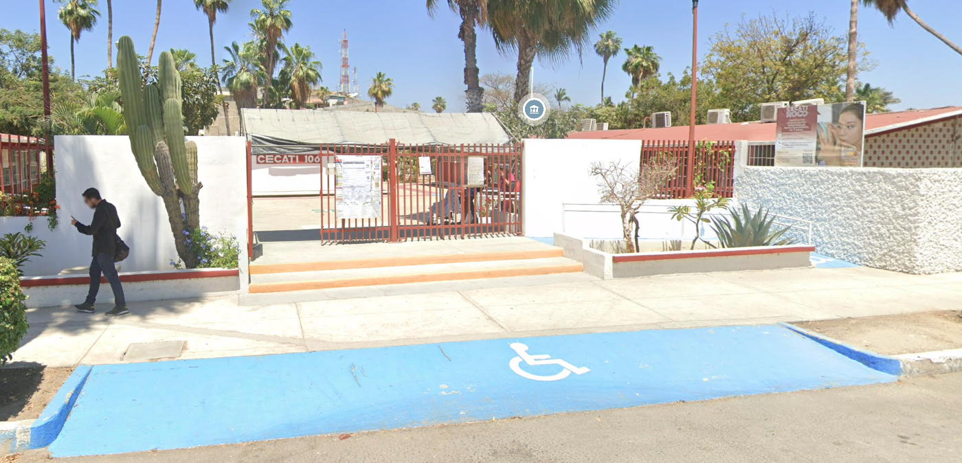 Fotografía en la que se muestra la entrada del plantel conformada por una reja de color rojo en medio de dos muros blancos y frente a ellos se encuentran dos jardineras. Al fondo hay un cartel que dice CECATI 106.